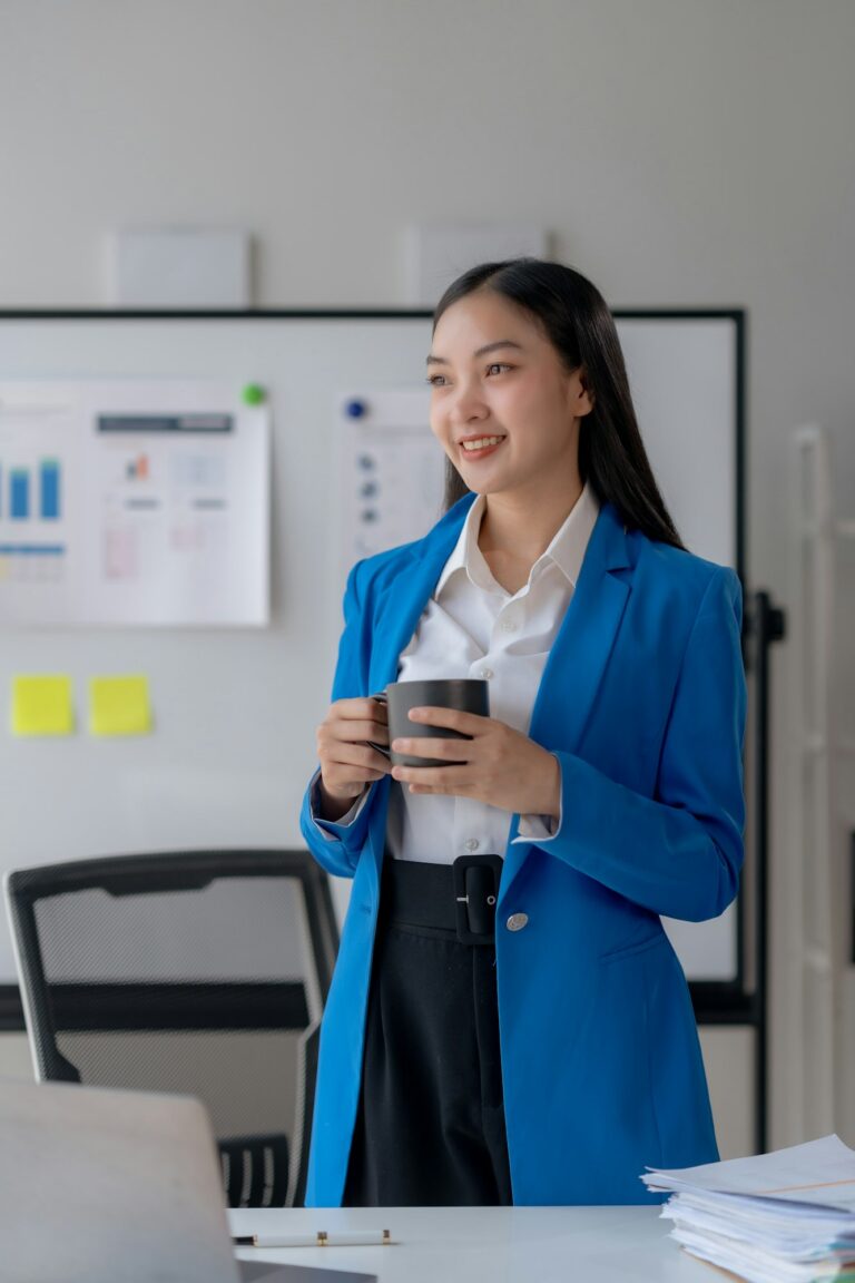 Confident young businesswoman in blue blazer enjoying coffee break at office desk